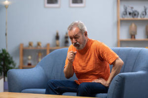An elderly man is sitting on a sofa at home while experiencing symptoms of rheumatoid arthritis.