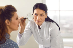 Dermatologist with magnifying glass looks at mole or pigmentation on woman's face.